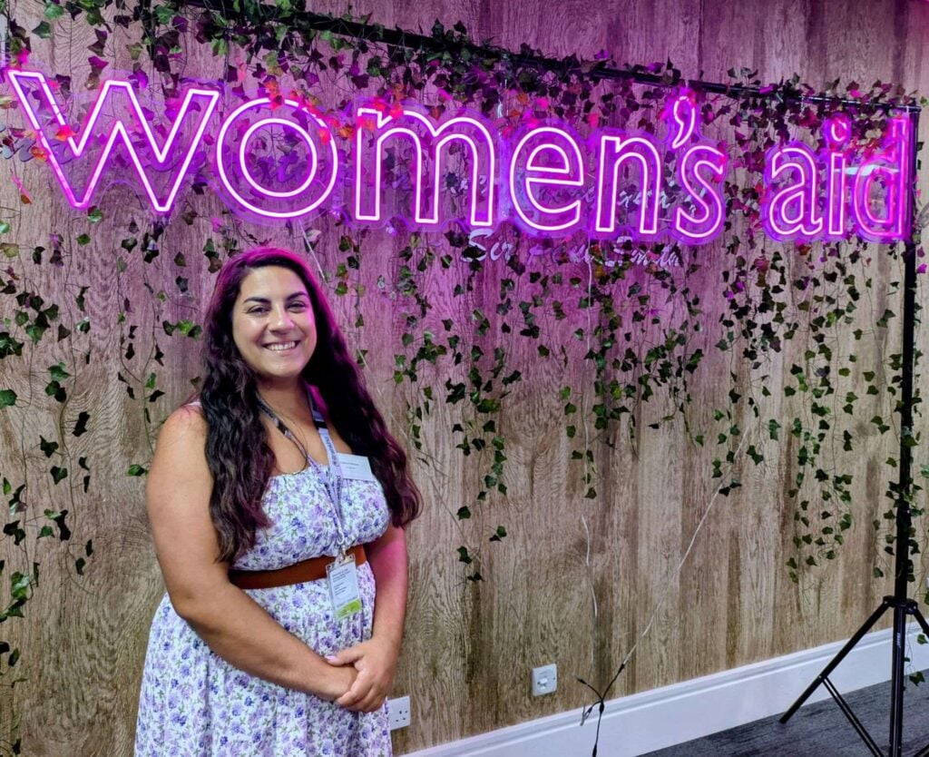 Steffy Bechelet, a woman with long hair and tanned skin, smiles at the camera. She stands in front of a neon sign reading 'Women's Aid'.