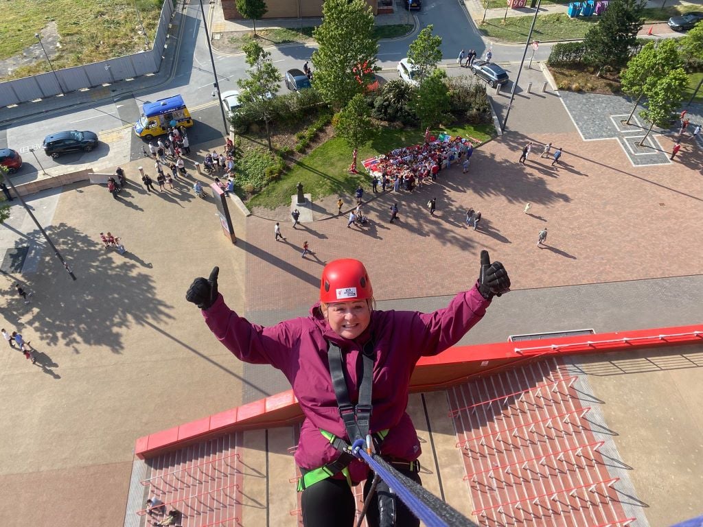 Julie leans backwards from a high wall, wearing a red helmet and with arms outstretched and thumbs up. In the background on the ground, a small crowd of people are cheering her on.