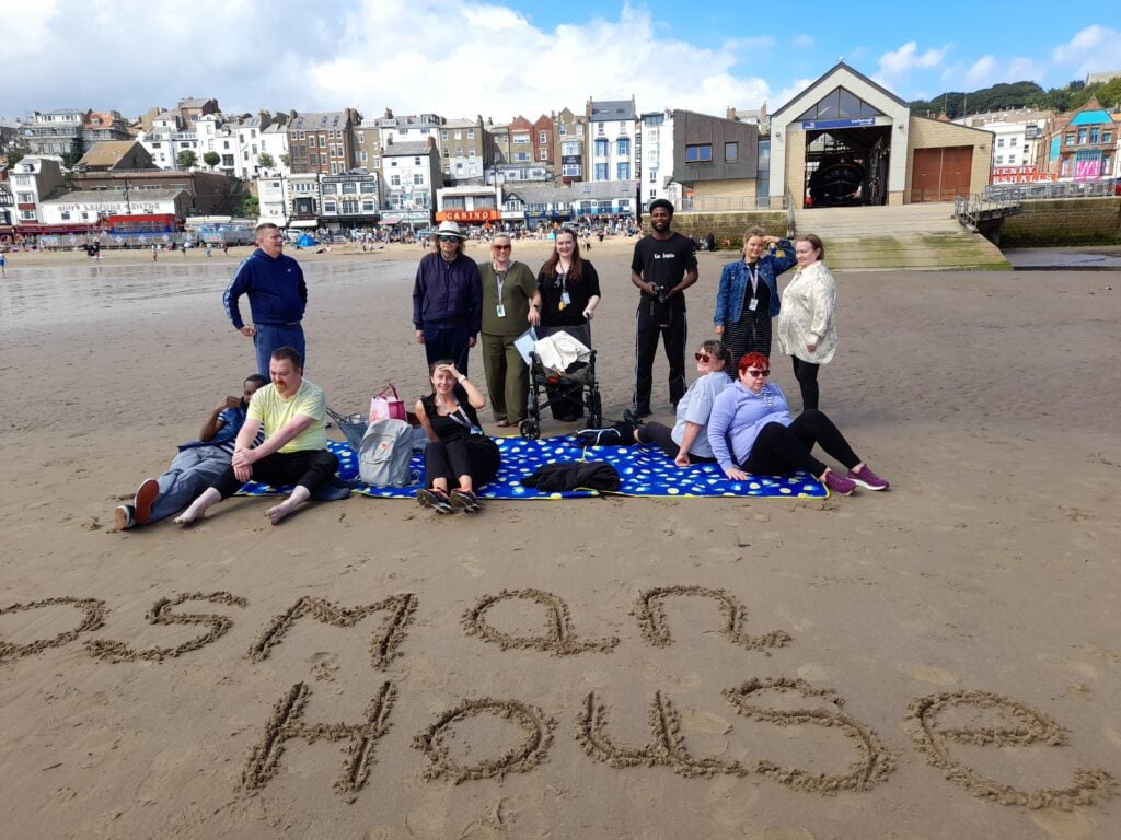 Five people are sitting on a blue picnic rug on the ground and seven people are standing behind them, are on a sandy beach at Scarborough, with the buildings on the seafront behind them. The words 'Osman House' are written in the sand in front of them.