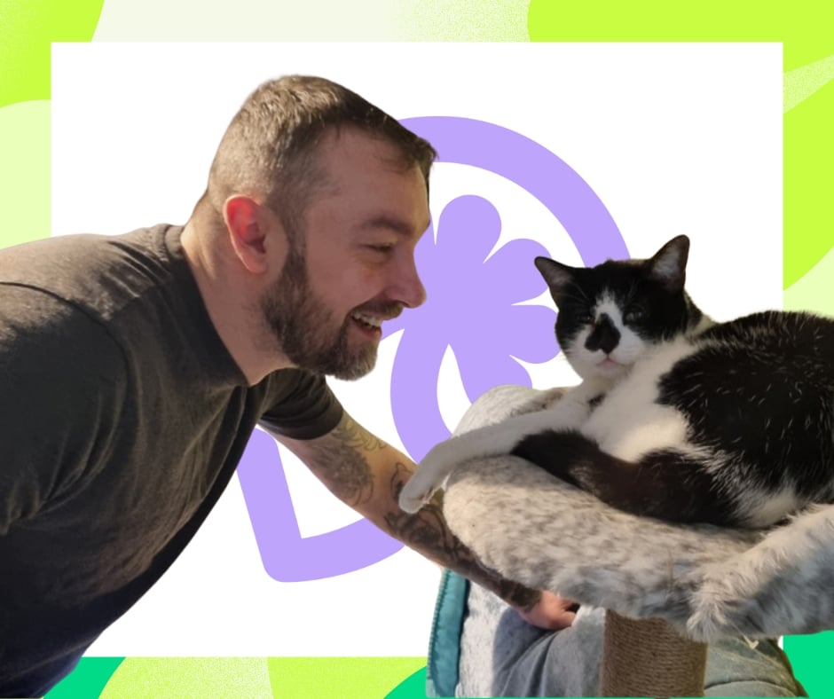 A photograph of Maciej, a white middle-aged man, leaning down to look at his black-and-white cat. He is smiling. The cat is sat on a cat-tree and looks relaxed.
