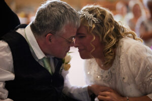 A photograph from Andy and Jane's wedding. Andy and Jane are touching foreheads with their eyes closed, happy. Andy is wearing a black suit and glasses. Jane is wearing a white wedding dress, shite shimmery headband, and has her hair in ringlets.