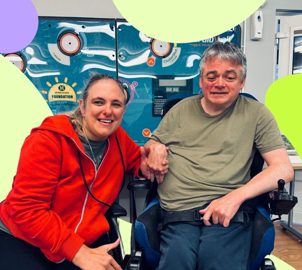 Jane and Andy are sitting down, holding hands and smiling at the camera. Jane is an adult white woman with brown hair in a ponytail. Andy is an adult white man with short greying hair. Andy is sat in his wheelchair. Andy and Jane are holding hands.